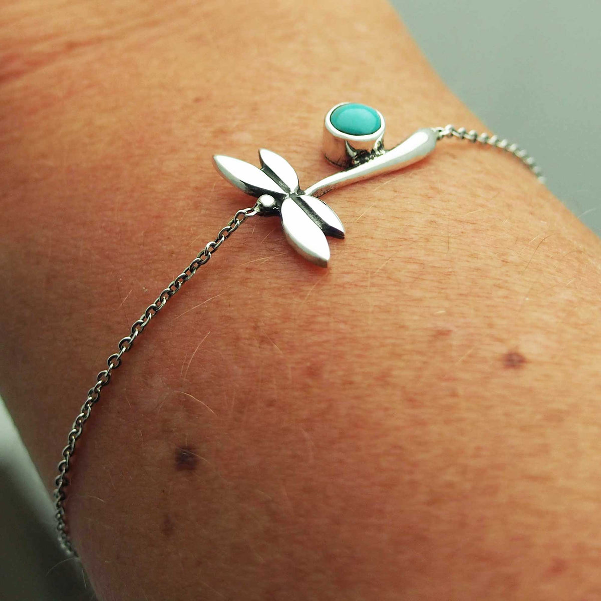 A silver bracelet with a dragonfly design and a turquoise stone on a person's wrist. Main image