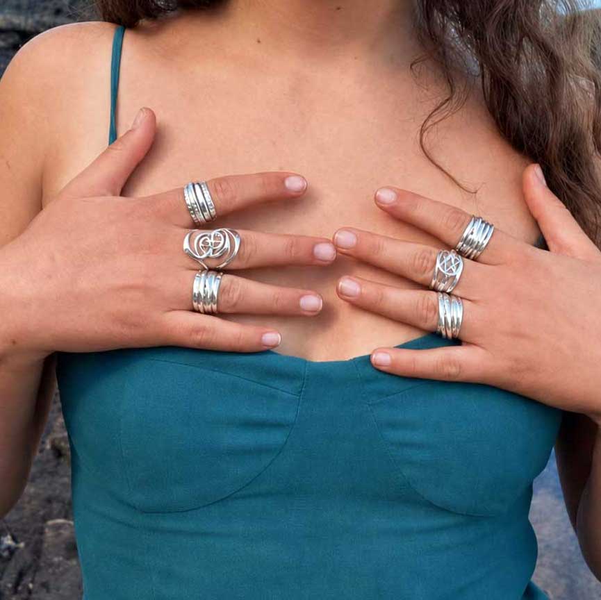 A close-up of a person wearing multiple silver rings on both hands, with the hands resting on the chest. The person is wearing a teal green top.