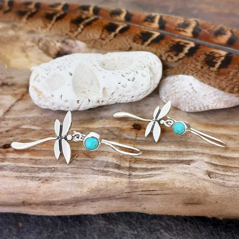 Dragonfly-shaped earrings with turquoise stones on a wooden surface with pebbles and feathers.