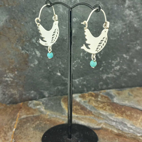 A pair of silver hoop earrings with cutout dove designs and small turquoise blue bead drops, displayed on a black stand.