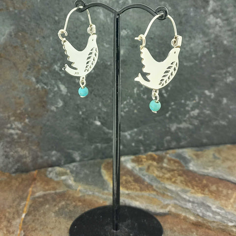 A pair of silver hoop earrings with cutout dove designs and small turquoise blue bead drops, displayed on a black stand.