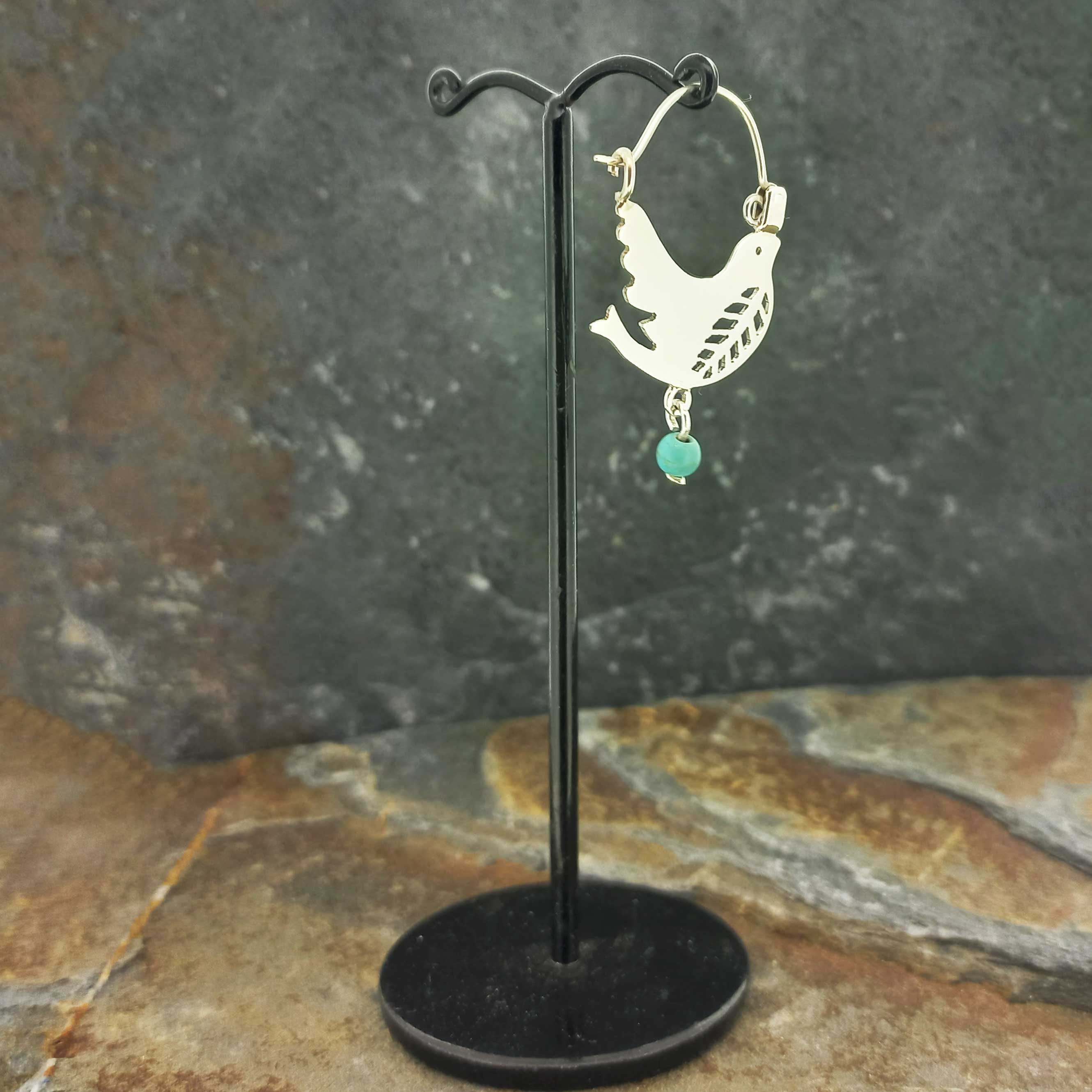 Silver hoop earrings with a flying dove cutout and a small turquoise bead dangle, displayed on a black stand.