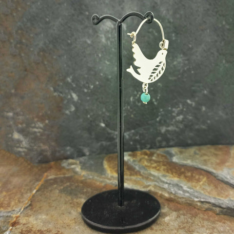 Silver hoop earrings with a flying dove cutout and a small turquoise bead dangle, displayed on a black stand.