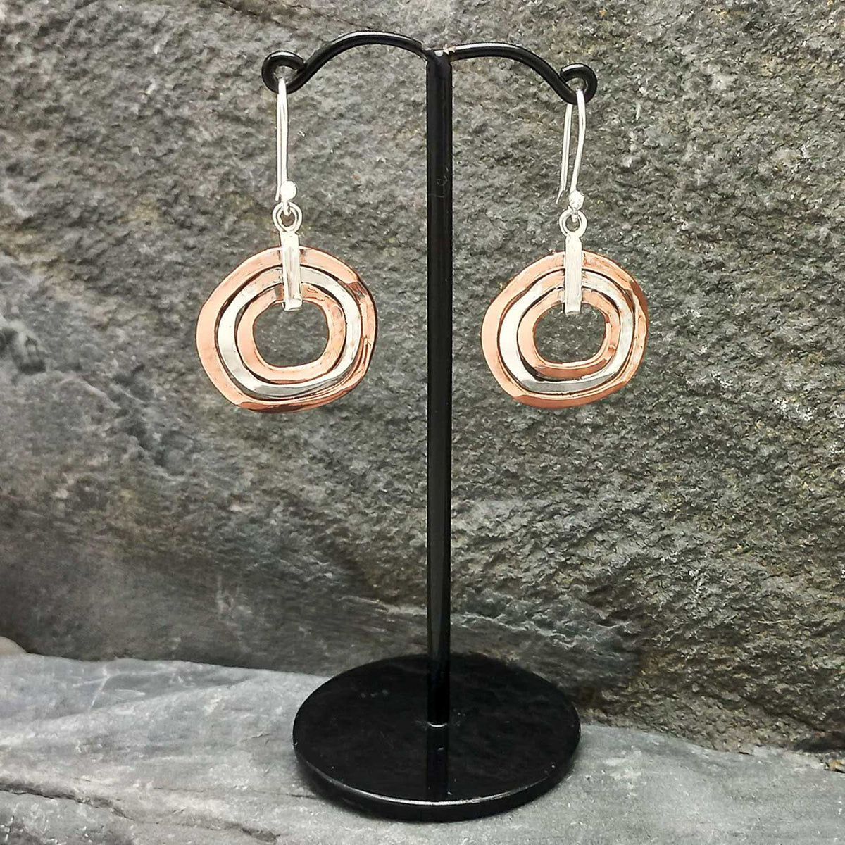 Pair of unusual copper silver circular earrings on a black stand against a stone background