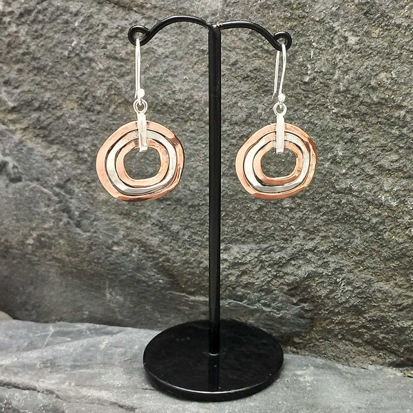Pair of unusual copper silver circular earrings on a black stand against a stone background