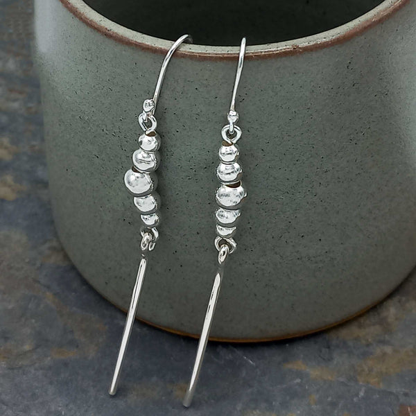 Silver beaded earrings on a textured surface with a ceramic pot in the background