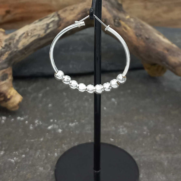 Silver hoop earring with bead detailing on a black stand against a natural wood and stone background