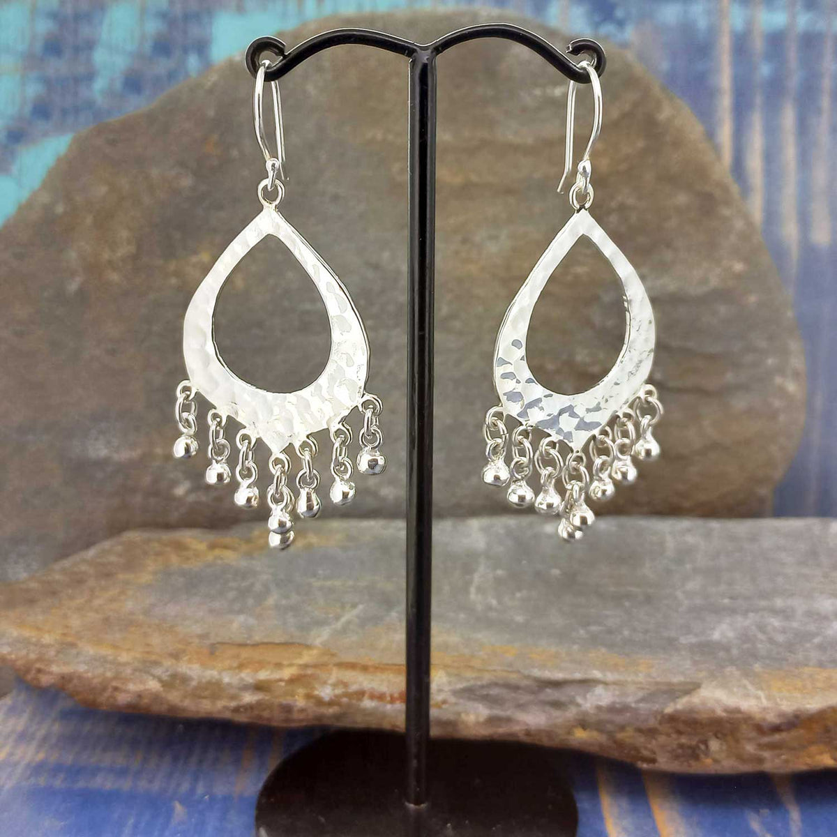 Silver teardrop Mexican earrings with dangling beads on a black stand against a stone background