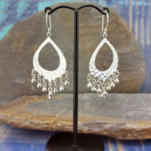 Silver teardrop Mexican earrings with dangling beads on a black stand against a stone background