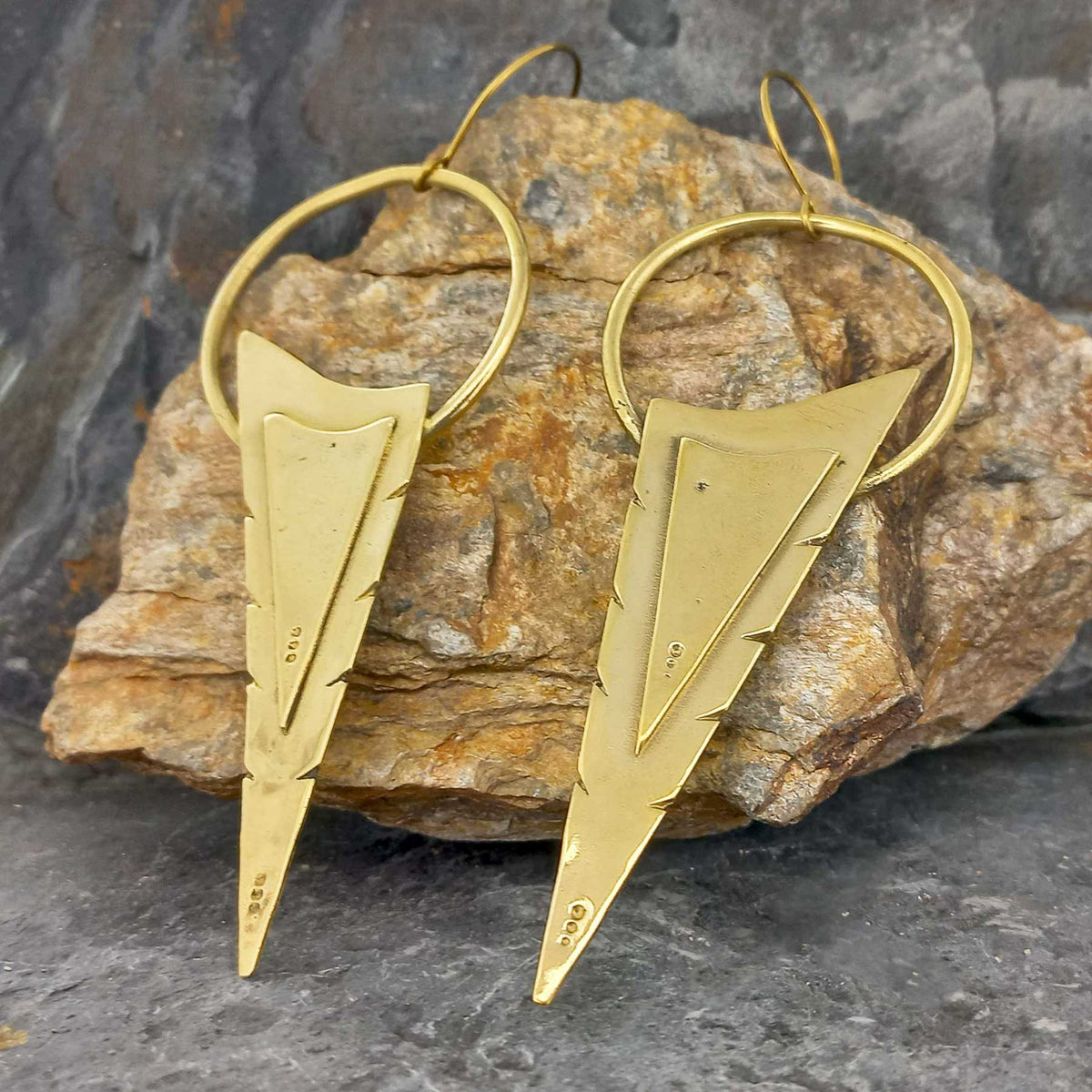 Gold coloured arrowhead-shaped brass earrings on a stone surface Main image