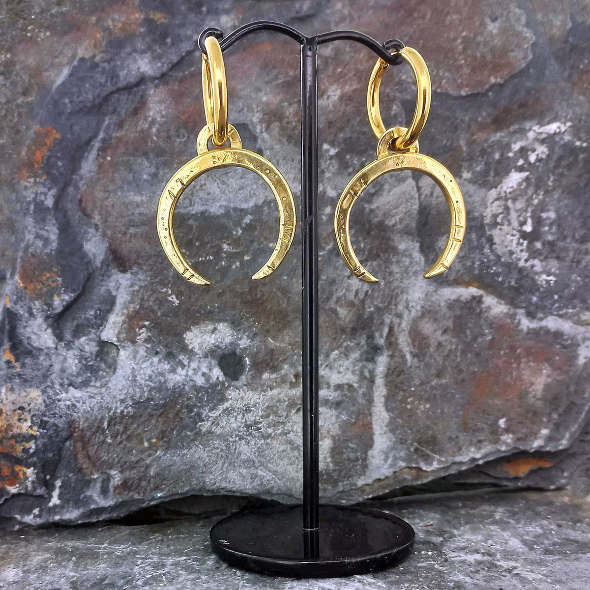 Gold hued brass crescent moon earrings on a black stand against a stone background