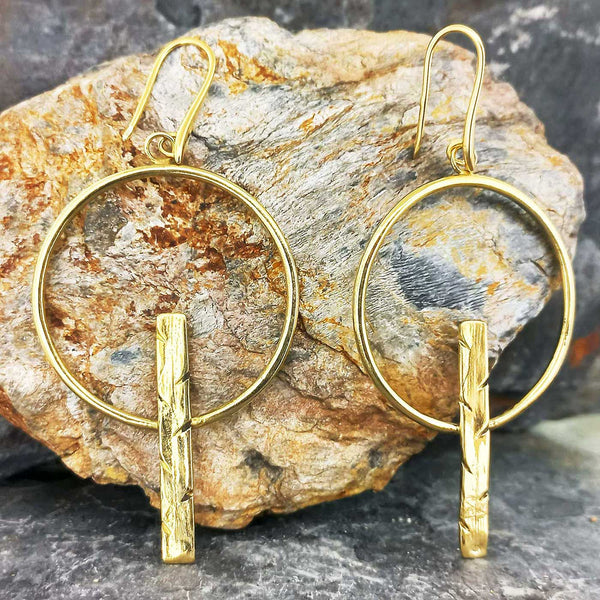 Gold coloured hoop earrings with vertical bars and gold plated hooks on a stone background