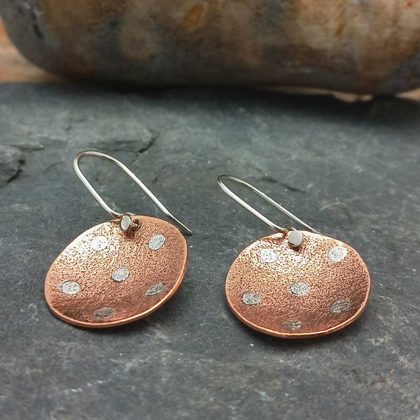Pair of copper earrings with textured surface and silver dots on a dark stone background