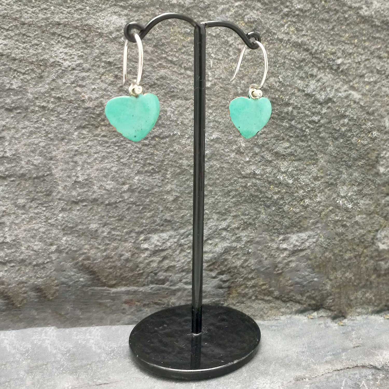 Turquoise heart-shaped earrings on a black stand against a stone wall.