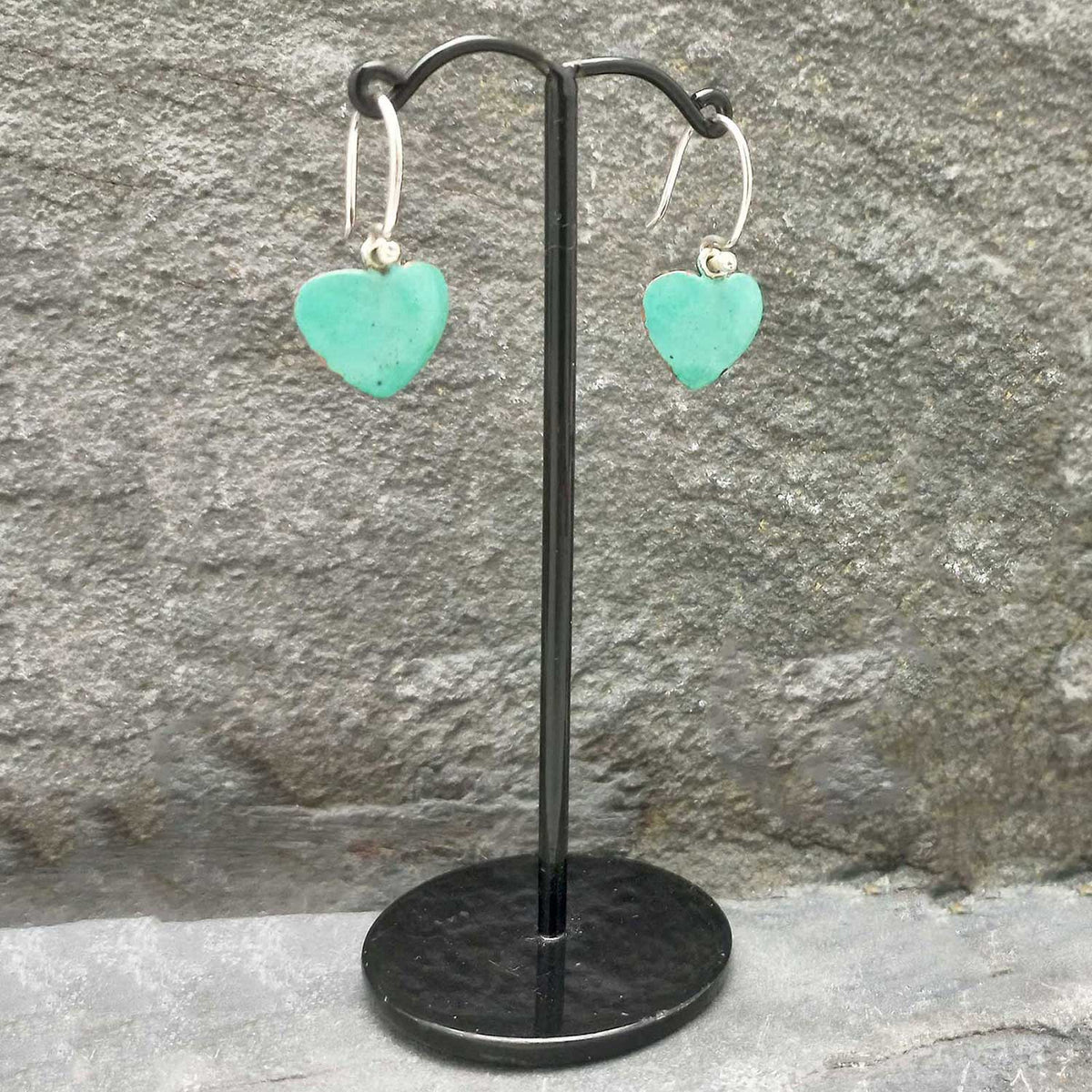 Turquoise heart-shaped earrings on a black stand against a stone wall.