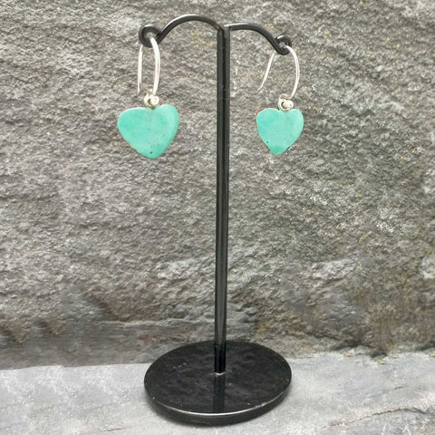 Turquoise heart-shaped earrings on a black stand against a stone wall.