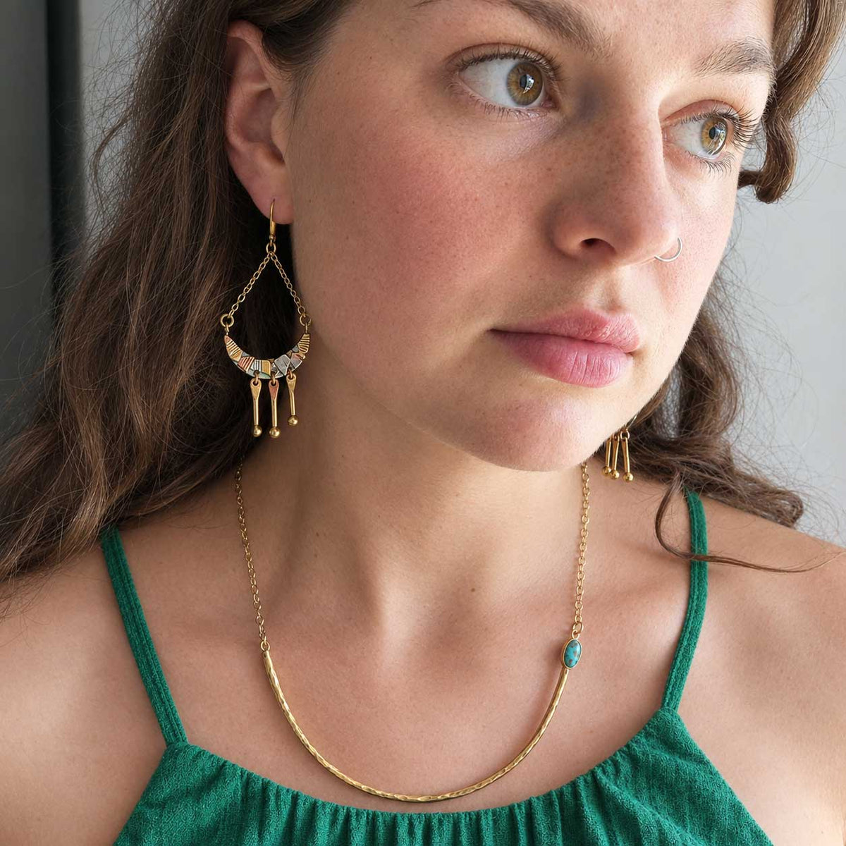 A close-up lifestyle shot of a woman wearing golden brass copper jewellery, featuring a crescent moon dangle earring and a delicate gold chain necklace with a turquoise accent.