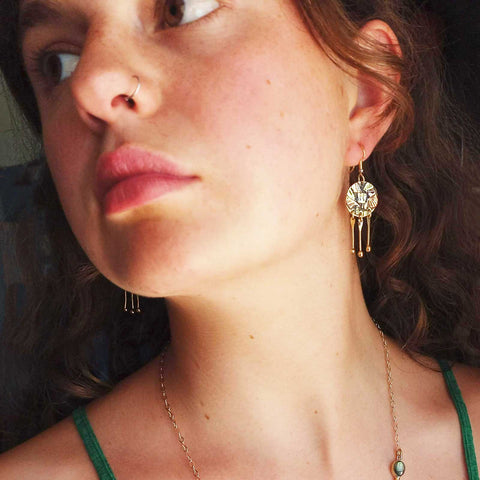 A close-up lifestyle shot of a woman wearing unusual gold toned earrings with dangling circular charm and three hanging loops.