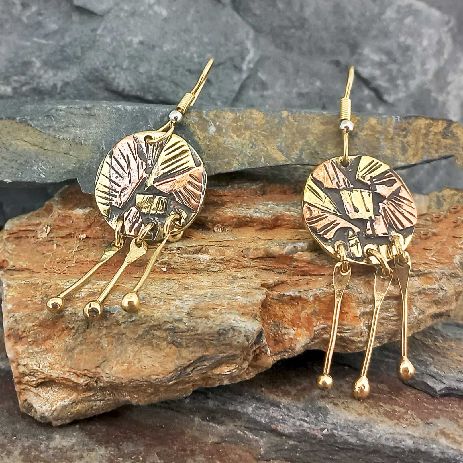 A pair of handmade brass and copper dangle earrings displayed on a natural rock surface. The earrings feature textured circular discs with engraved details and three dangling bar charms with small gold beads.