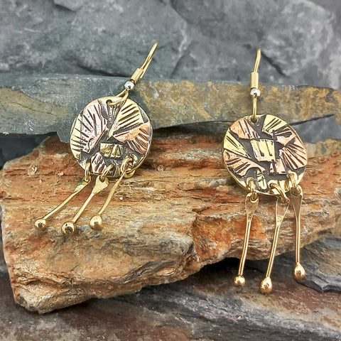 A pair of handmade brass and copper dangle earrings displayed on a natural rock surface. The earrings feature textured circular discs with engraved details and three dangling bar charms with small gold beads.