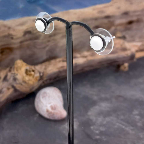 Silver oxidised circle stud earrings on a black stand with a natural background of driftwood and a shell.