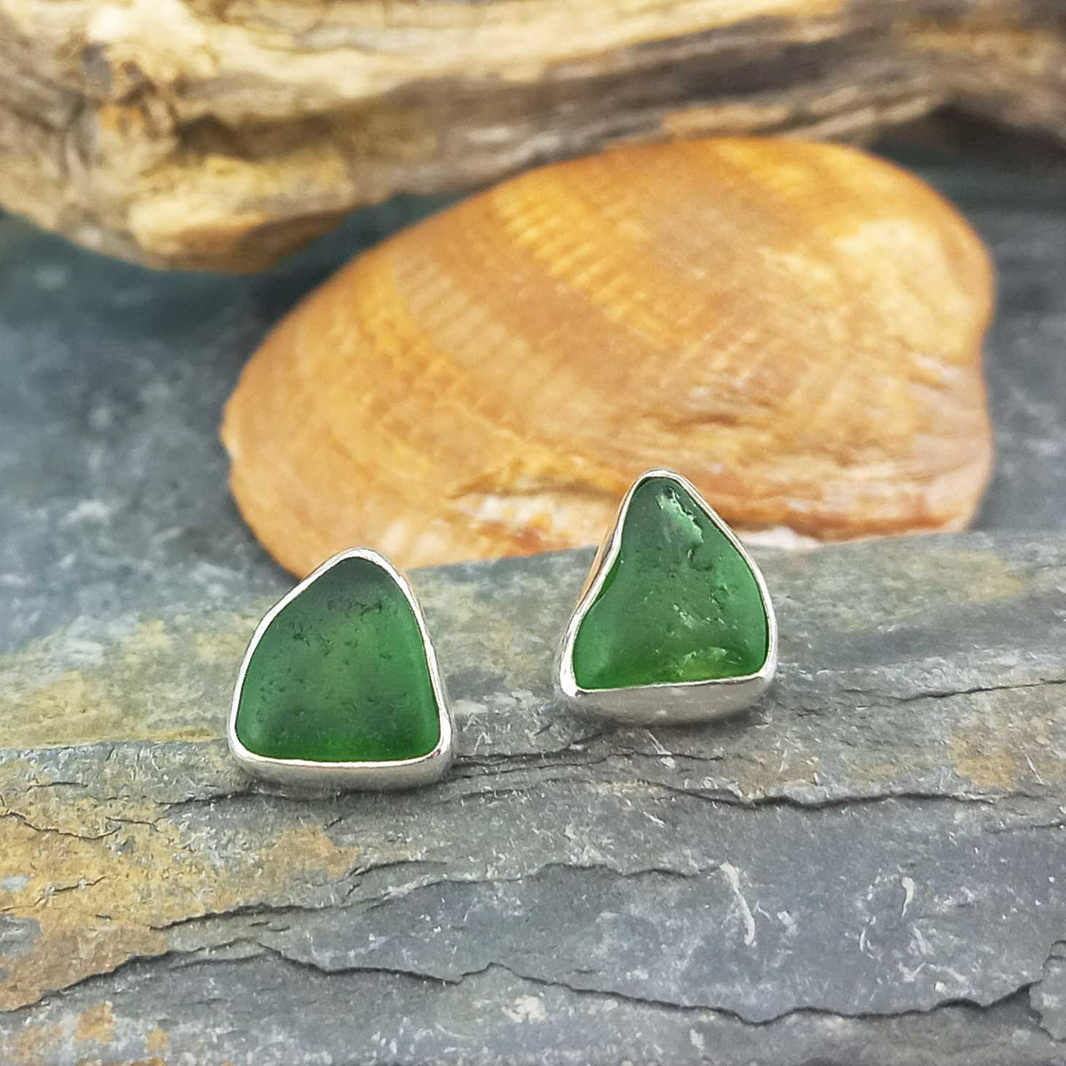 A pair of triangular sea glass stud earrings with vibrant green stones set in silver, displayed on a slate surface with a seashell and driftwood in the background.