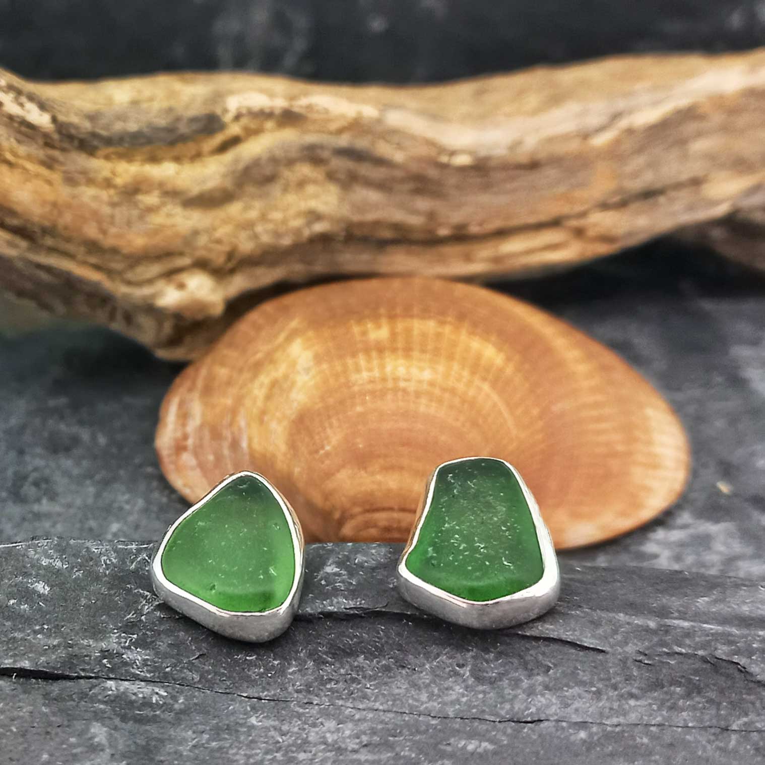 Pair of handmade sea glass stud earrings with green stones in sterling silver bezel settings, displayed on a dark textured surface with a clam shell and driftwood in the background.