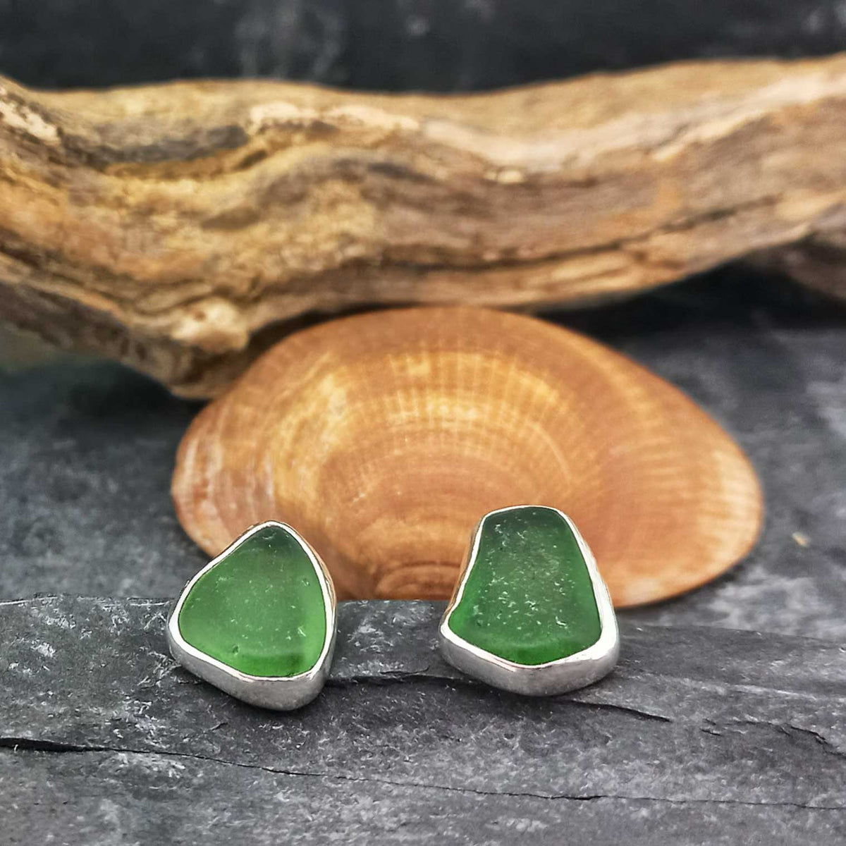 Pair of handmade sea glass stud earrings with green stones in sterling silver bezel settings, displayed on a dark textured surface with a clam shell and driftwood in the background.