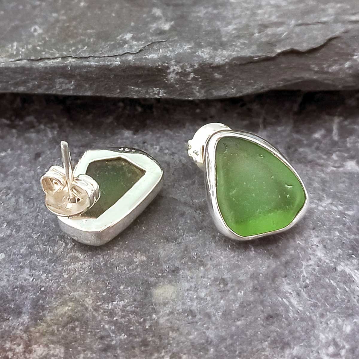 Two handmade sea glass stud earrings with green stones in sterling silver bezel settings, one shown from the side and the other from the back with the post visible, resting on a gray slate surface.