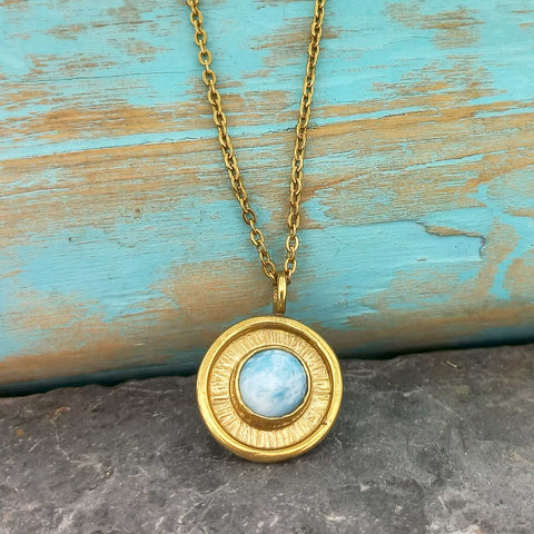 Gold coloured necklace with a circular pendant featuring a blue stone on a textured surface.