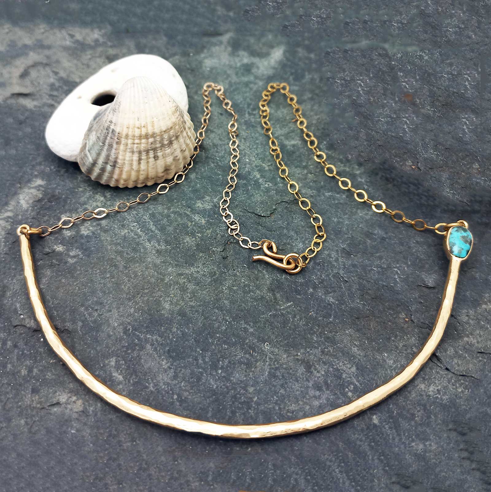 Unusual gold-tone necklace with a hammered curved bar, a delicate chain, and a small turquoise-blue stone.