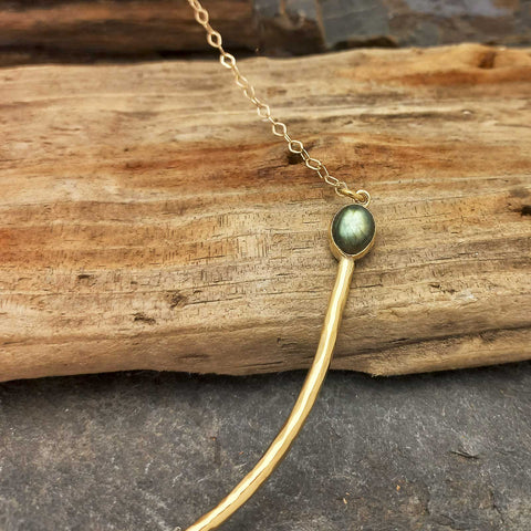 A brass necklace with a curved bar pendant and a green Labradorite stone, displayed on a piece of weathered wood.