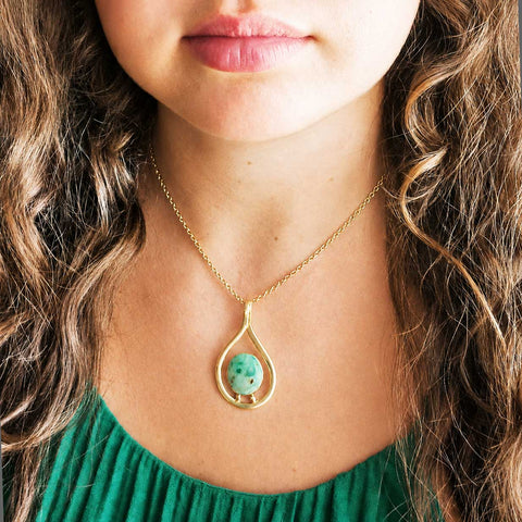 A close-up lifestyle shot of a woman wearing a gold teardrop pendant necklace with a green marbled Chrysocolla stone centrepiece, paired with a green top.