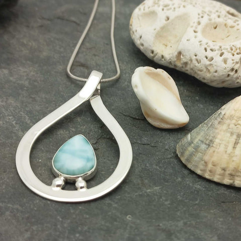 A sterling silver teardrop pendant necklace with a light blue Larimar stone, displayed on a dark slate surface next to seashells.