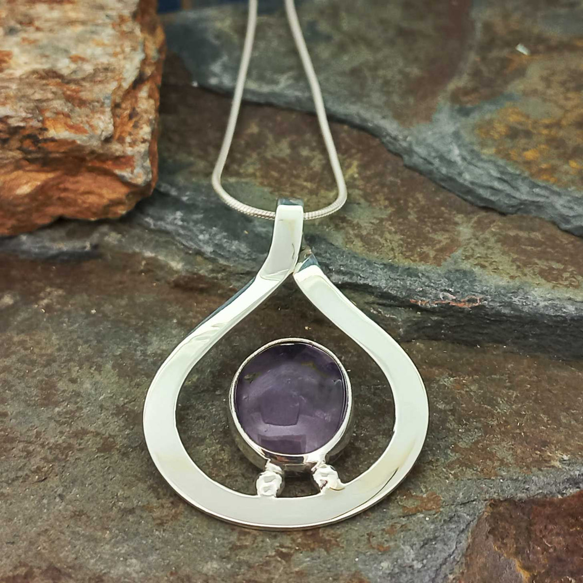 A silver teardrop pendant necklace with a bezel-set purple amethyst stone, displayed on a textured slate surface.