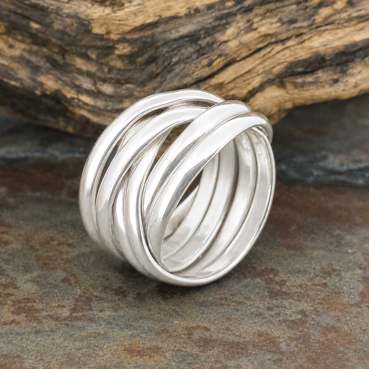 A wide chunky polished silver ring with multiple interlocking bands, resting on a dark stone surface with a piece of weathered driftwood in the background.