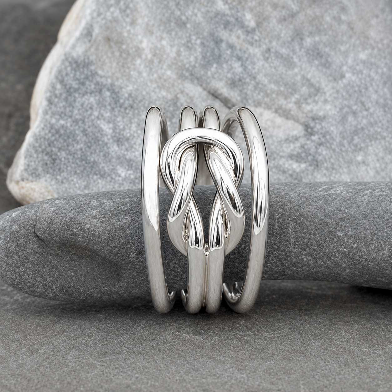 A polished sterling silver ring with a central knotted design and three parallel bands, displayed on a smooth gray stone.