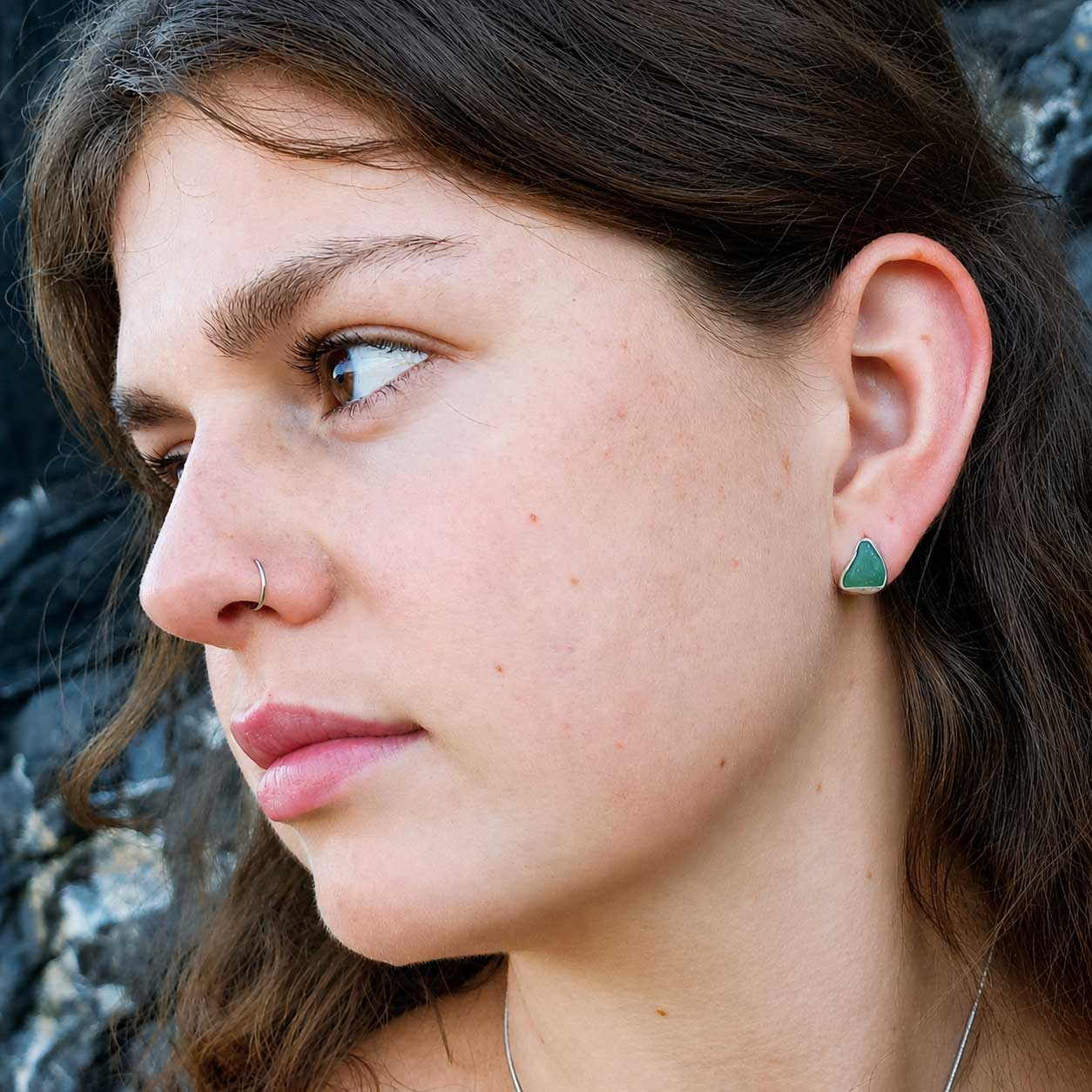 A close-up side profile of a woman outdoors wearing a small triangular green sea glass stud earring.
