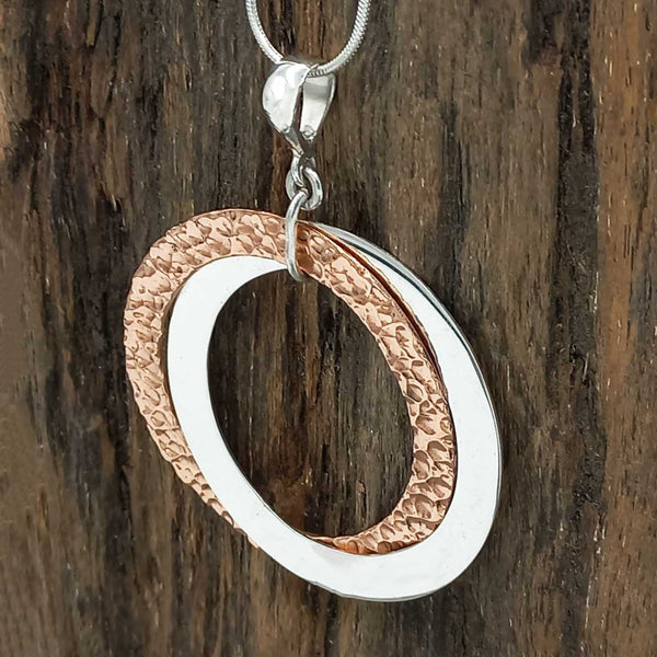 Jewelry pendant with two interlocking rings, one silver and one copper, on a wooden background.
